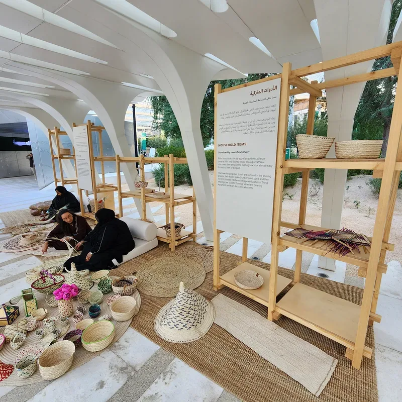 Household items exhibit featuring handwoven palm leaf baskets, fans, and mats under arched canopy