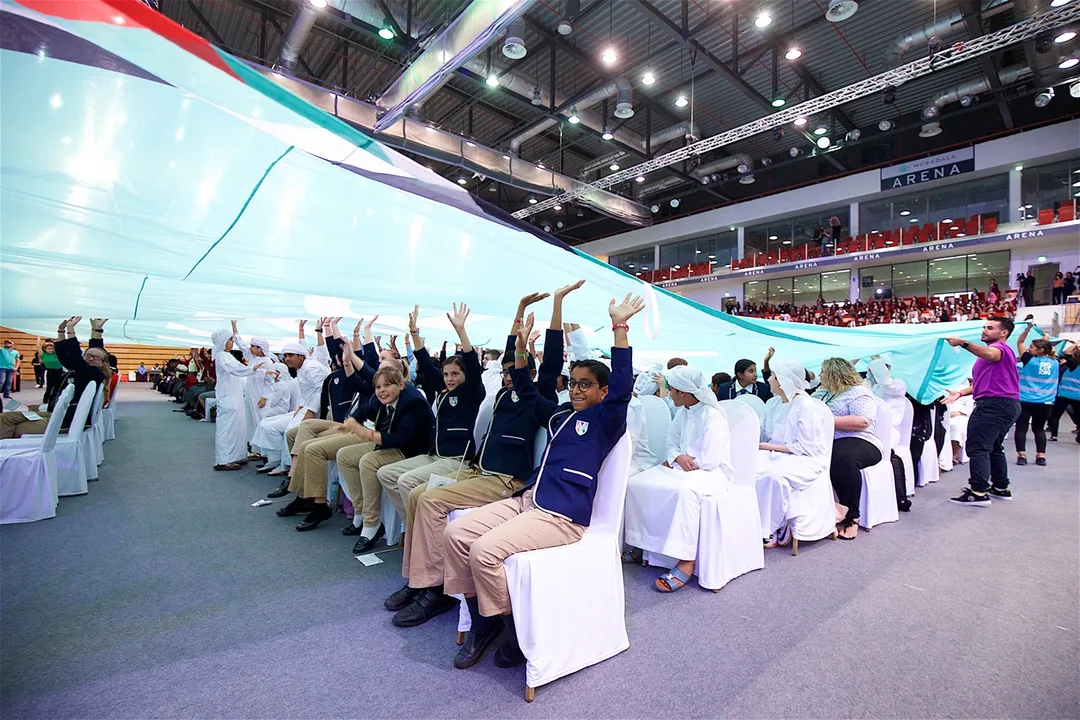 Youth Tolerance Rally participants lifting a large UAE flag above seated students inside Mubadala Arena.