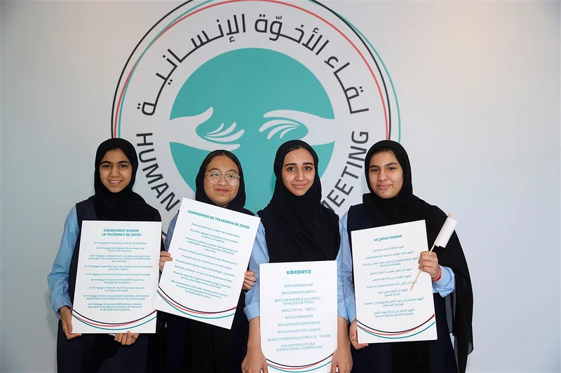 Four students holding multilingual tolerance pledges in front of the Human Fraternity Meeting logo